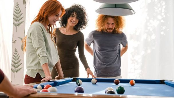 週末のパーティー中に自宅で明るいゲームルームのプールテーブルでビリヤードボールを見ている幸せな多民族男性と女性の友人 - 写真・画像