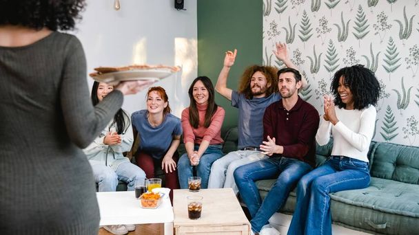 Разнообразная группа друзей-мужчин и женщин, болеющих за анонимную женщину, приносящую пиццу в гостиную на выходных, собираются вместе дома - Фото, изображение