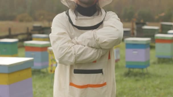Tilt up medium slowmo portret van Kaukasische mannelijke imker in beschermende kleding poseren met de handen gevouwen op bijenstal met kleurrijke houten bijenkorven op de achtergrond - Video