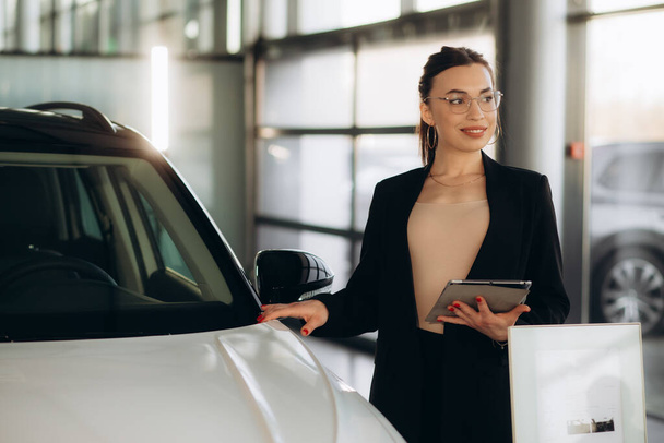Smiling saleswoman holding tablet PC while looking at camera at new car showroom. Professional car dealer posing proudly at auto showroom, smiling to the camera, holding tablet PC. - Photo, Image