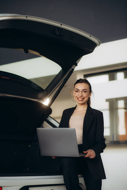 Venditore femminile sorridente in giacca e cravatta utilizzando tablet per guardare quale auto viene venduta mentre in piedi nel salone di auto. - Foto, immagini