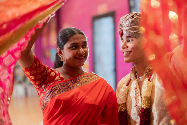 Una mujer en un sari y un hombre en un sherwani ríen juntos, rodeados de vibrantes cortinas. - Foto, imagen