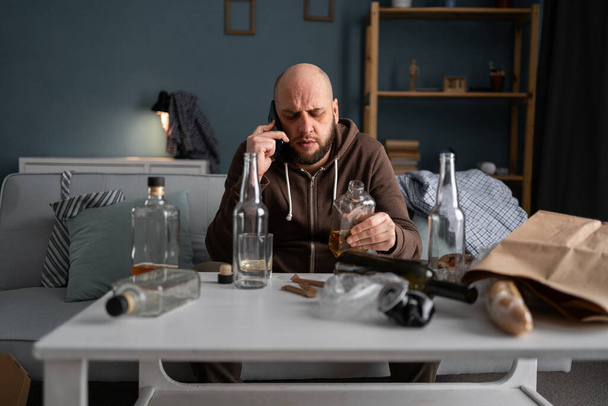Részeg férfi otthon whiskys üveget tart alkoholizmussal, alkoholfüggőséggel és drogfüggőséggel. Hívás mobilon, rendetlen szobában. - Fotó, kép