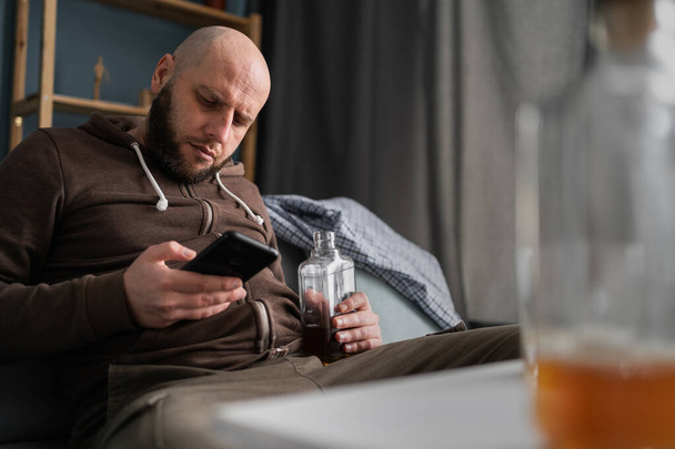 Adicción al alcohol y concepto de personas. Hombre borracho con teléfono inteligente y botella de whisky sentado en el sofá en casa. Copiar espacio - Foto, Imagen