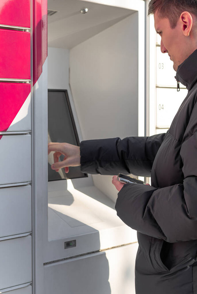 One young Caucasian handsome man in a dark jacket is holding a mobile phone in his hand, and the other is entering information on the screen to receive his parcel from the box, standing on the street - Photo, Image