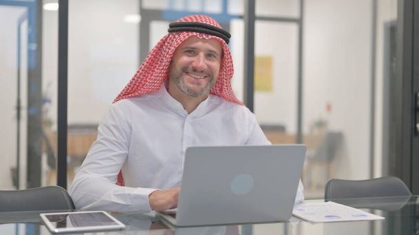 Arab Man uśmiecha się do kamery podczas pracy na laptopie w biurze - Zdjęcie, obraz