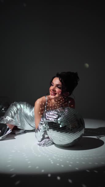 Woman in silver lying beside disco balls. Joyful woman with disco ball in silver attire. Gleaming Joy at the Disco - Footage, Video