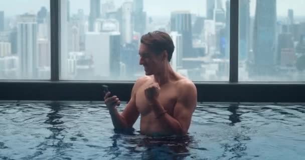 Hombre rico usando smartphone y relajarse en la piscina de la azotea de rascacielos en la gran ciudad. Millennial turista masculino con smartphone en la mano en la azotea piscina de hotel de lujo con fondo de ciudad. - Metraje, vídeo