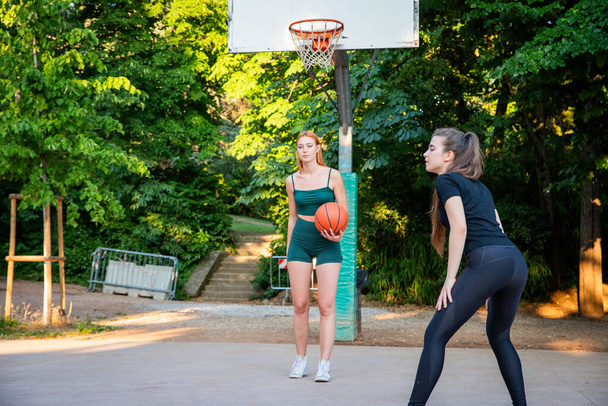 Δύο γυναίκες που παίζουν μπάσκετ σε ένα πάρκο. Ένας από αυτούς κρατάει μια μπάλα του μπάσκετ. Η άλλη γυναίκα φοράει πράσινο μπλουζάκι. - Φωτογραφία, εικόνα