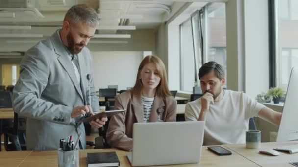 Jonge zakenvrouw en haar mannelijke collega 's bespreken project op laptop en digitale tablet terwijl ze samenwerken in modern open ruimte kantoor - Video