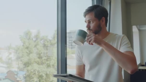 Молодий чоловік дивиться через вікно в офісі, п'є каву з чашки і працює на цифровому планшеті - Кадри, відео