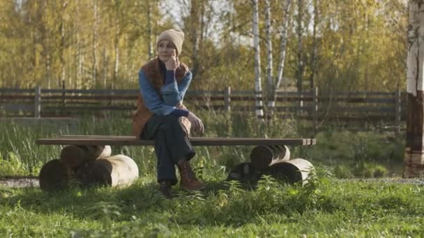 Plně pomalý portrét středně dospělé bělošky sedící na dřevěné lavičce na venkově se slunečním světlem a hledící do kamery - Záběry, video