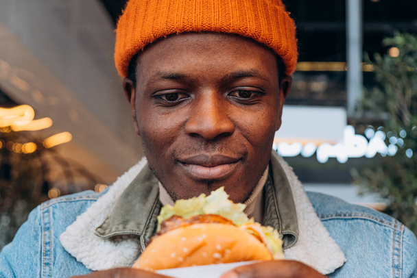 Afro-Amerikaanse Man in Beanie Holding en Kijkend naar een Heerlijke Burger met Anticipatie. Een close-up van een man met een warme glimlach, klaar om te genieten van een sappige hamburger, gevangen in een levendige stedelijke eetgelegenheid - Foto, afbeelding