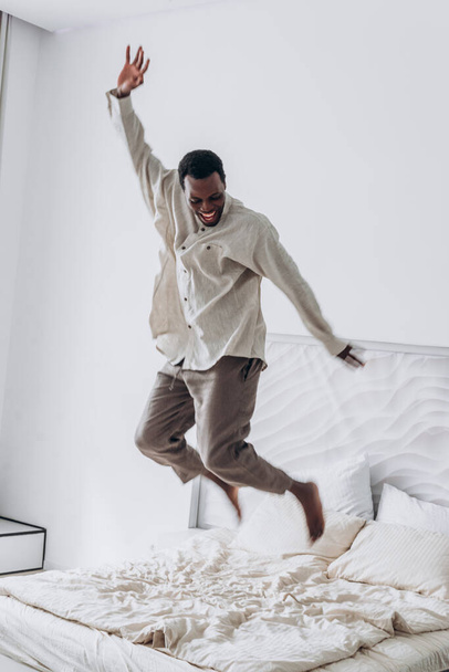 Hombre afroamericano alegre saltando en la cama en el dormitorio brillante. Un hombre exultante saltando sobre una cama, brazos levantados en un momento despreocupado, su risa y movimiento encapsulando pura alegría en un dormitorio luminoso - Foto, imagen