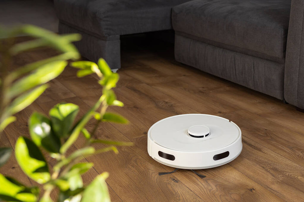 A fehér robot porszívó tisztítja fa laminált a padlón a nappaliban. port gyűjt a kanapé közelében nehéz helyeken és leveleket a nagy kanapé alól. infravörös érzékelők és kamerák vezérlése. - Fotó, kép
