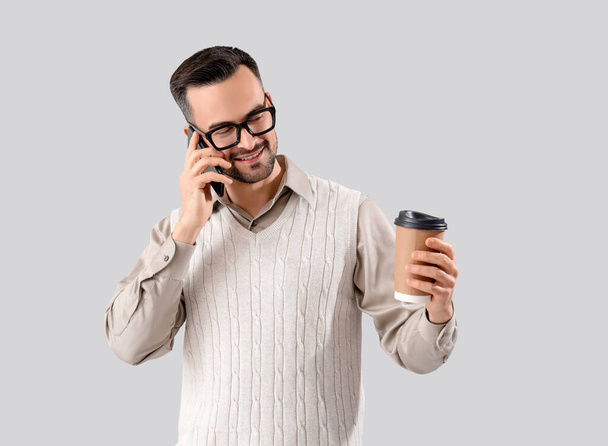 Schöner Mann mit Brille und Kaffeetasse im Gespräch per Handy auf hellem Hintergrund - Foto, Bild