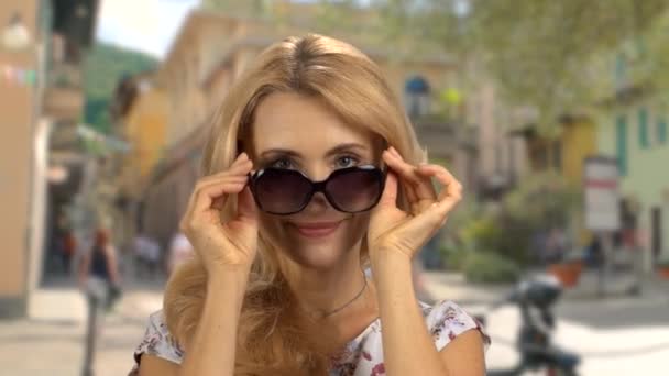 Portret van een blonde vrouw die op straat staat en een zonnebril opzet. Buiten op de achtergrond. - Video