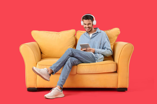 Guapo joven feliz hombre en los auriculares con la tableta descansando en el sofá sobre el fondo rojo - Foto, imagen