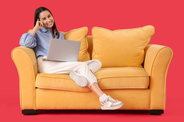 Schöne junge glückliche Frau mit Laptop ruht auf Sofa vor rotem Hintergrund - Foto, Bild