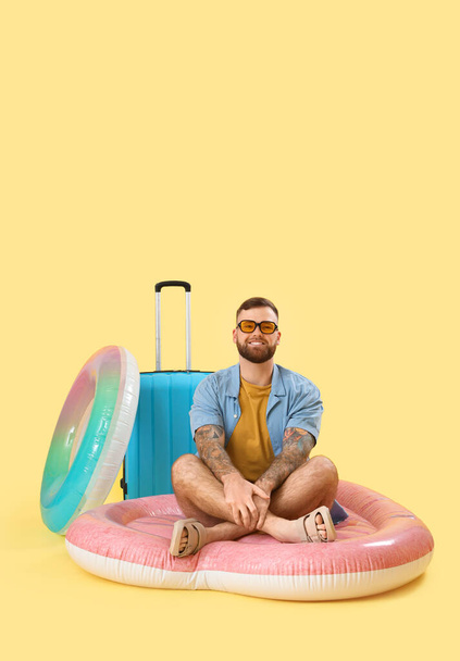 Молодой человек с надувным кольцом, матрасом и чемоданом на желтом фоне - Фото, изображение
