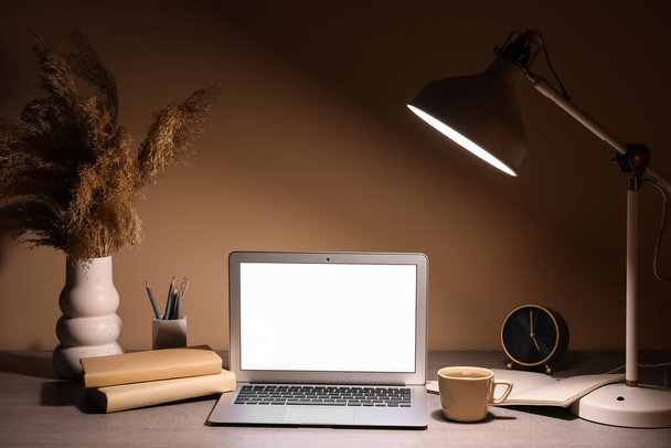 Local de trabalho moderno com lâmpada brilhante, laptop, copo e relógio à noite - Foto, Imagem