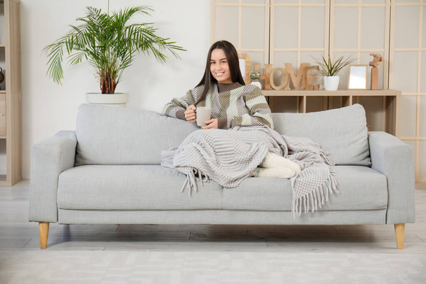 Kaunis nuori nainen kupin teetä ja lämmin huopa istuu harmaa sohva olohuoneessa - Valokuva, kuva