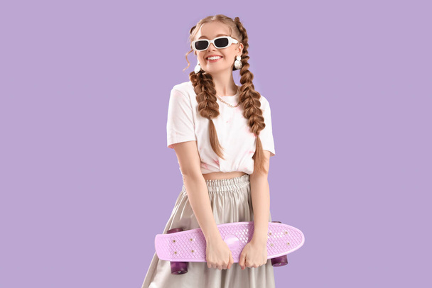 Glücklich lächelnde junge Frau mit Zöpfen in Sonnenbrille und Skateboard auf fliederfarbenem Hintergrund - Foto, Bild