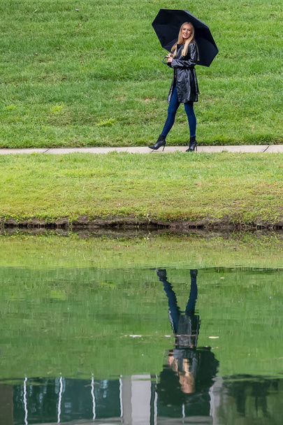 Una mujer impresionante pasea por el parque, su elegancia acentuada por un paraguas negro, en medio de la penumbra que se avecina antes de que la lluvia bese la tierra. - Foto, Imagen