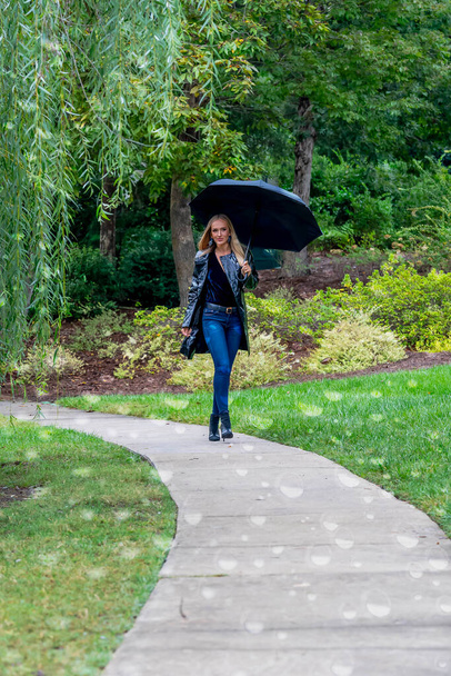 Una mujer impresionante pasea por el parque, su elegancia acentuada por un paraguas negro, en medio de la penumbra que se avecina antes de que la lluvia bese la tierra. - Foto, Imagen