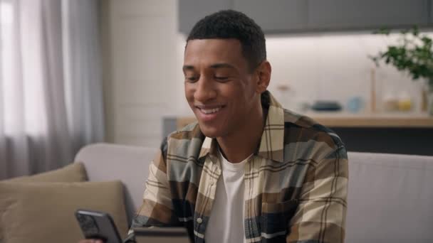 Afro-Américain acheteur utilisateur insouciant gars ethnique mâle client souriant heureux excité ordre livraison en ligne app bancaire paiement réservation achats avec carte bancaire de crédit et téléphone mobile à la maison - Séquence, vidéo