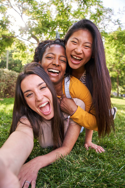 Pystysuora selfie ryhmä monirotuinen iloinen nuori innoissaan naiset poseeraavat nauraa makaa päällekkäin ruohoa puistossa. Kolme onnellista naista katselee kameraa nauttien kesälomasta yhdessä ulkona - Valokuva, kuva