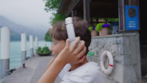 Glücklicher kleiner Junge hört Musik, schüttelt den Kopf im Takt des Liedes, mit Blick auf den See und die Berge in einer wunderschönen Landschaft - Filmmaterial, Video