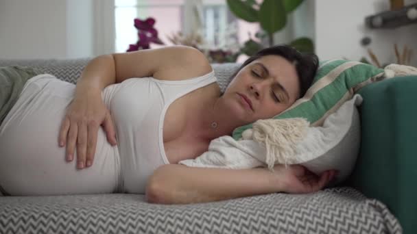 Ciężarna kobieta odczuwa dyskomfort w późnym stadium ciąży, kobieta po trzydziestce próbuje odpocząć w domu na kanapie zmaga się z nudnościami - Materiał filmowy, wideo