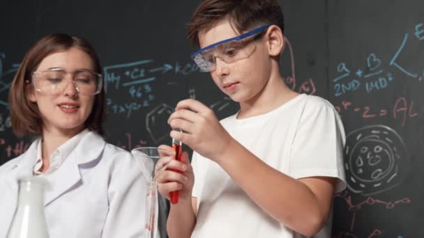 Slimme jongen giet chemische vloeistof in het bekerglas terwijl de leraar geeft advies op STEM science class. Student mengen gekleurde oplossing tijdens het staan aan boord geschreven chemie theorie in het laboratorium. Erutie. - Video