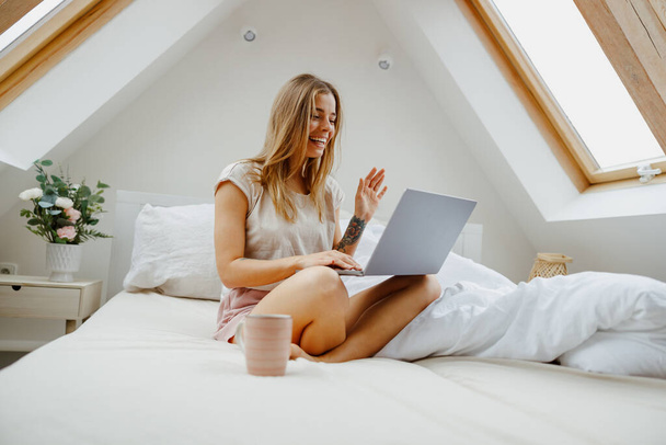 Una mujer está cómodamente sentada en una cama, usando su computadora portátil con sus dedos - Foto, imagen