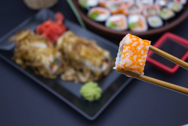 Tämä kiehtova kuva tuo kokemus sushi ruokailu elämään lähikuva sushi pala hallussa bambu syömäpuikot, keskittynyt etualalla, jossa hämärtynyt tausta valikoituja sushi rullina lautasella. Kirkas oranssi Masago s - Valokuva, kuva