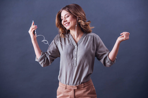 Ελκυστική νεαρή γυναίκα στο smart casual ρούχα και τα ακουστικά χρησιμοποιώντας ένα smartphone, να ακούτε μουσική και να χαμογελά, σε γκρίζο φόντο - Φωτογραφία, εικόνα