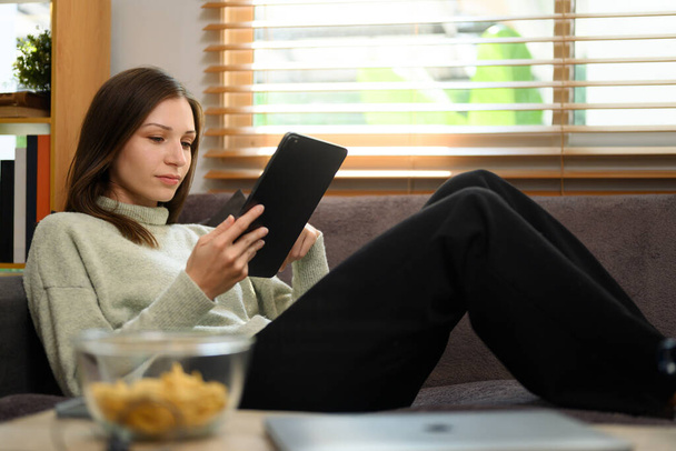 Młoda piękna kobieta za pomocą tabletu cyfrowego na kanapie w salonie. - Zdjęcie, obraz