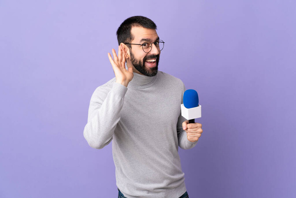 Homem repórter adulto com barba segurando um microfone sobre fundo roxo isolado ouvindo algo, colocando a mão na orelha - Foto, Imagem