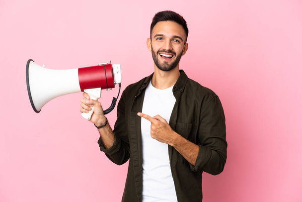 Young caucasian man isolated on pink background holding a megaphone and pointing side - Photo, Image