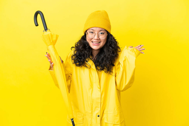 Donna asiatica con impermeabile e ombrello isolato su sfondo giallo con espressione facciale scioccata - Foto, immagini