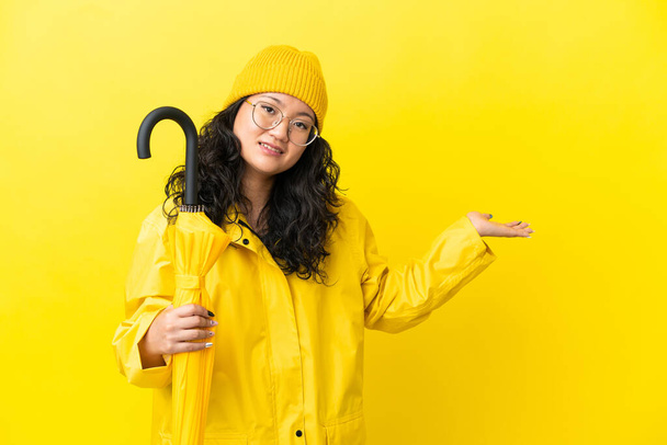 Donna asiatica con impermeabile e ombrello isolato su sfondo giallo che estende le mani di lato per invitare a venire - Foto, immagini
