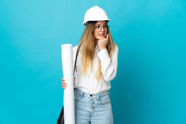 Jeune architecte femme avec casque et tenant des plans isolés sur fond bleu ayant des doutes et la pensée - Photo, image