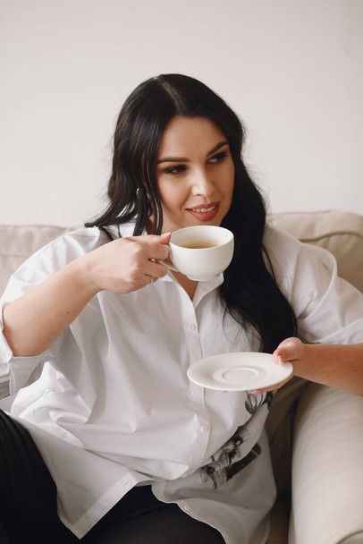 Portrait of smiling brunette woman in white shirt enjoying a cup of coffee at home. Relax time. Peaceful lady having relaxing day, chilling on lazy morning - Photo, Image