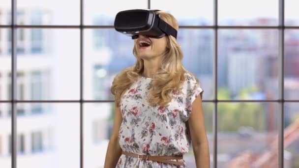 Happy blonde woman wears vr headset indoors. Checkered window with cityscape view in the background. - Footage, Video