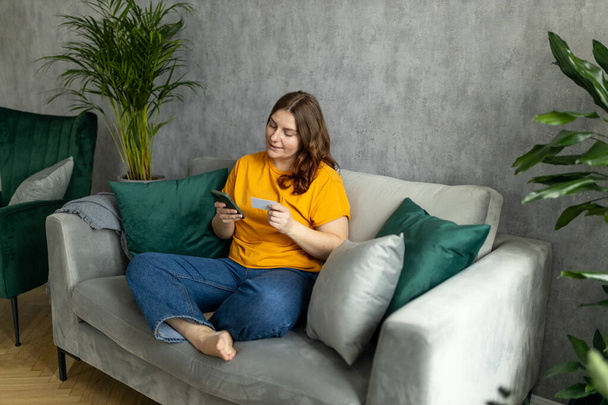 Cheerful young woman in casual clothes sit on couch spending time in living room at home. Rest relax good mood leisure lifestyle concept. 30s female Using mobile phone, hold credit bank card - Foto, Imagen