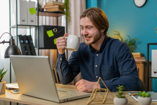 Young Caucasian businessman working on laptop computer drinking cup of warm hot coffee sits at desk workplace home office room in the morning. Male freelancer guy enjoying relaxing, taking a break. - Photo, Image