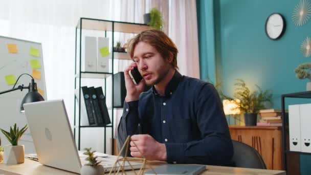 Young businessman freelancer answering to client on mobile phone call, using smartphone and laptop. Conversation. Caucasian man guy working online remote distance job, talking at home office desk. - Footage, Video