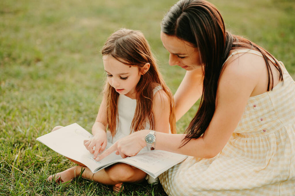 mooie jonge moeder besteden tijd met haar dochter lezen in park terwijl zitten op het gras. - Foto, afbeelding