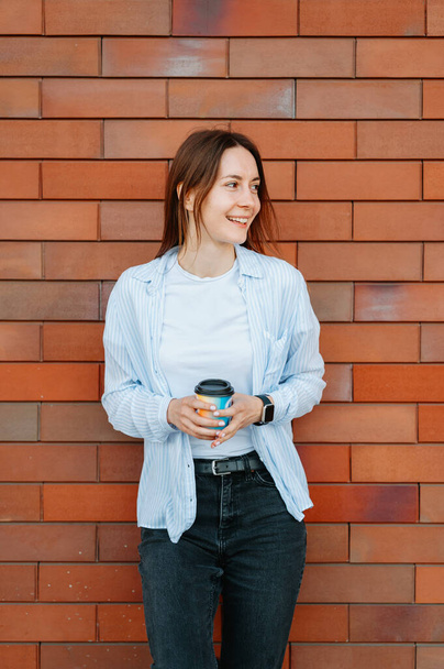 Вертикальный снимок веселой молодой женщины, держащей чашку кофе у кирпичной стены. - Фото, изображение